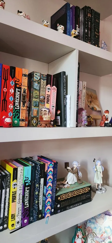 Estante branca com livros de mitologia e folclore, entre bonecos e miniaturas